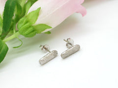 A pair of silver earrings with a textured design rests on a white surface, accompanied by a delicate pink flower.