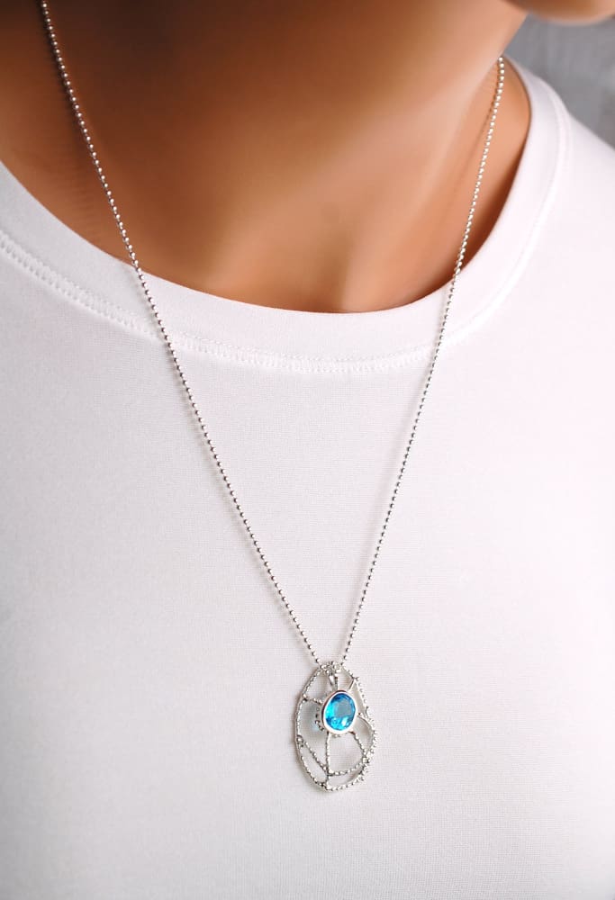 A silver necklace with a blue gemstone pendant is worn by a person in a white shirt.