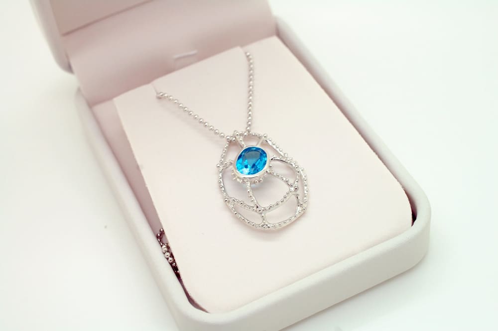 A silver necklace with a blue gemstone pendant is displayed in an open jewelry box.