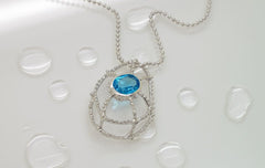 A silver necklace with a blue gemstone pendant is displayed on a white surface with water droplets scattered around it.